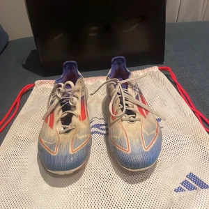 Adidas F50 elite Fg - Snygga Adidas fotbollsskor i vitt med blå och röda detaljer. Skorna har snörning och är perfekta för fotbollsspel på gräs. De har en modern design med en kombination av färger som ger ett sportigt utseende.