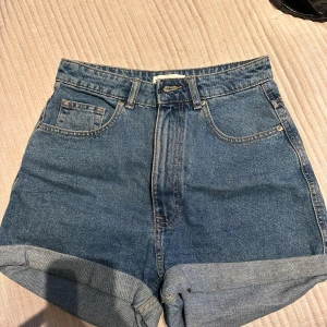 Blå jeansshorts från Zara - Snygga blå jeansshorts från Zara med hög midja och klassisk femficksdesign. Perfekta för sommardagar med en avslappnad stil. De har en uppvikt kant och stängs med dragkedja och knapp.