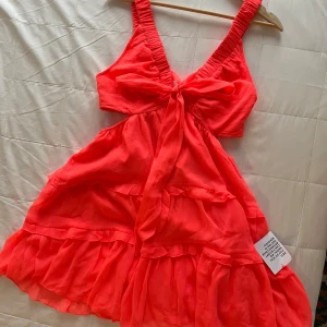 Korallfärgad klänning med volanger - Snygg korallfärgad klänning med volanger och knytband framtill. Den har en v-ringad design och breda axelband. Perfekt för sommarens alla tillfällen!