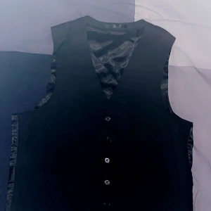 Svart väst med knappar - Elegant svart väst med knappar framtill och justerbart spänne bak. Perfekt för en stilren look.