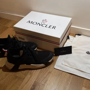 Svarta skor  Moncler - Snygga svarta sneakers från Moncler med vit sula och Monclers ikoniska röd-vit-blå detalj på hälen. Skorna har snörning och en modern design som passar perfekt för en stilren look. Köpta i NK