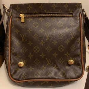 Brun axelväska med monogram - Snygg brun axelväska med klassiskt monogrammönster och justerbar axelrem. Väskan har en dragkedja framtill och dekorativa metalldetaljer. Perfekt för att bära dina dagliga nödvändigheter med stil. 24x24x5 cm