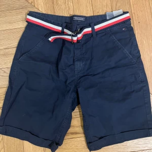 Mörkblå shorts från Tommy Hilfiger - Snygga mörkblå shorts från Tommy Hilfiger med ett stilrent bälte i rött, vitt och blått. Perfekta för sommardagar med en klassisk design och bekväm passform. Shortsen har praktiska fickor och en diskret logga.