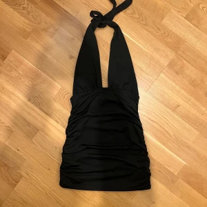 Halterneck klänning mini - Snygg svart halterneck klänning som knyts i nacken. Den är rynkad längst sidorna som ger en snygg passform längst kroppen och halva ryggen är öppen. Klänningen är kort och går lite under rumpan. Storlek 36/S och materialet är stretchigt. Passar perfekt till fester! Skriv gärna om du har några frågor🤗