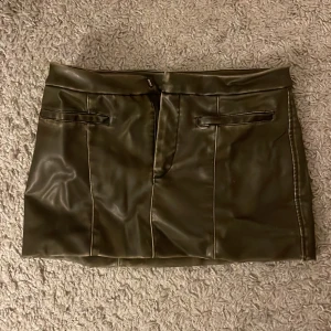 Olivgrön skinnkjol från Zara - Snygg olivgrön skinnkjol från Zara i kort modell. Kjolen har en stilren design med dragkedja bak och dekorativa fickor fram. Perfekt för att ge din outfit en edgy touch. Köparen står för frakt!