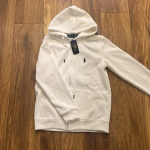 Vit hoodie från Ralph Lauren - Säljer en stilren vit hoodie från Ralph Lauren med dragkedja och den klassiska loggan på bröstet. Perfekt för en avslappnad look. Sitter som en S men står M i tröjan. Splitter ny