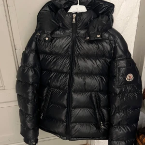 Marinblå dunjacka från Moncler - Snygg marinblå dunjacka från Moncler med huva och dragkedja. Jackan har långa ärmar och en logotyp på ärmen. Perfekt för kyliga dagar. Självklart äkta. Finns äkta hets bevis och en bag till själva jackan.