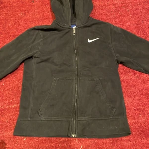 Svart hoodie från Nike - Säljer en svart hoodie från Nike med dragkedja och huva. Den har en vit Nike-logga på bröstet och praktiska fickor framtill. Perfekt för en sportig look eller avslappnade dagar.