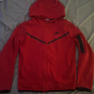 Röd huvtröja från Nike - Säljer en röd huvtröja från Nike med dragkedja och svarta detaljer. Perfekt för en sportig look med långärmade ärmar och en bekväm passform.