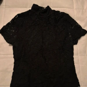 Svart spetsblus med dragkedja - Elegant svart spetsblus med korta ärmar och hög krage. Blusen har en dragkedja i ryggen för en stilren look. Perfekt för en chic och sofistikerad stil. (Lånade bilder)