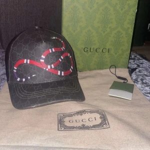 Gucci keps  - Snygg svart keps från Gucci med ett rött och vitt ormtryck. Kepsen har justerbar rem baktill med röd detalj och är tillverkad i ett slitstarkt material med Gucci-logotypen diskret mönstrad över hela ytan.