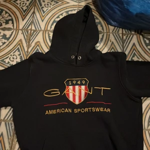 Svart hoodie från Gant - Säljer en svart hoodie från Gant med ett stort tryck på framsidan som inkluderar texten 'American Sportswear' och ett emblem. Hoodien har en klassisk design med dragsko i huvan.