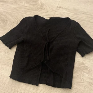 Svart ribbad kofta från Lindex - Säljer en svart ribbad kofta från Lindex i storlek 146/152. Koftan har korta ärmar och knytning framtill, perfekt för en stilren look. Passar bra till både vardag och speciella tillfällen.