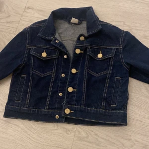Mörkblå jeansjacka från Lindex Kids - Säljer en snygg mörkblå jeansjacka från Lindex Kids. Jackan har klassisk design med knappar framtill och två bröstfickor med lock. Perfekt för vår och höst!
