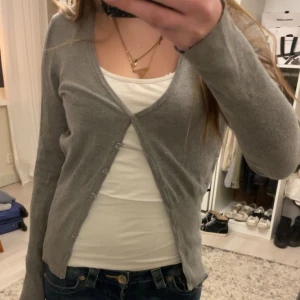 Grå kofta - Säljer en stilren grå kofta med knappar framtill. Perfekt för lager-på-lager och passar till många olika outfits. Koftan har långa ärmar och en v-ringad design.