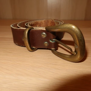Brunt skärp med mässingsspänne - Säljer ett stilrent brunt skärp i skinn med ett robust mässingsspänne. Perfekt för att ge en klassisk touch till din outfit. Skärpet har en enkel design med nitar vid spännet.