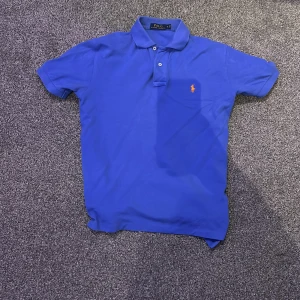 Blå pikétröja från Ralph Lauren - Snygg blå pikétröja från Ralph Lauren med klassisk krage och knappslå. Den har en liten orange logotyp broderad på bröstet. Perfekt för en stilren look.