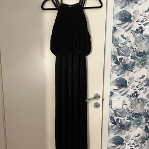 Svart långklänning med halterneck - Elegant svart långklänning med halterneck-design. Klänningen har en stilren och flytande siluett som passar perfekt för festliga tillfällen. Den är ärmlös och har en vacker drapering som ger en sofistikerad look. Rekommenderars som balklänning även.