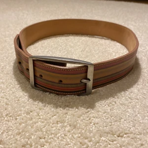 Randigt bälte från Paul Smith - Snyggt bälte från Paul Smith i skinn med ett randigt mönster i olika färger. Bältet har ett stilrent metallspänne och är perfekt för att ge en färgklick till din outfit.