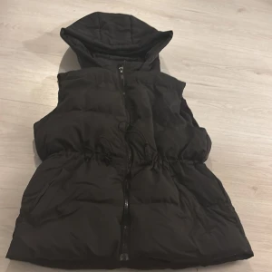 Svart dunväst med huva - Snygg svart dunväst med dragkedja och huva. Perfekt för kyliga dagar och ger en sportig look. Västen har en justerbar dragsko i midjan för bättre passform.