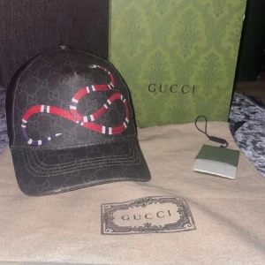 Svart keps från Gucci med ormmotiv - Snygg svart keps från Gucci med ett iögonfallande rött och vitt ormmotiv. Kepsen har justerbar passform och är tillverkad i Italien. Perfekt för att ge din outfit en lyxig touch.