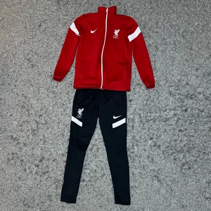 Liverpool tracksuit från Nike - Snygg tracksuit från Nike med vita detaljer och dragkedja. Jackan har klubbmärke på bröstet och Nike loggan och långa ärmar. Byxorna kommer också med vita detaljer med klubbmärket och Nike loggan.Perfekt för fotbollsträning eller som en sportig vardagslook.