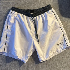 Vita shorts från Hugo Boss - Snygga vita shorts från Hugo Boss med mörkgrön resårmidja och snörning. Perfekta för sommardagar vid stranden eller poolen. Diskret logga på benet och praktisk bakficka.