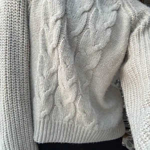 Beige stickad tröja med flätmönster - Mysig beige stickad tröja med ett snyggt flätmönster på framsidan. Tröjan har långa ärmar och ribbstickade kanter som ger en klassisk look. Perfekt för kyliga dagar.