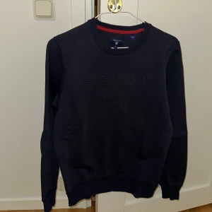 Mörkblå tröja från GANT - Säljer en stilren mörkblå tröja från GANT med broderad logotyp på bröstet. Tröjan har rund halsringning med en röd detalj på insidan av kragen och långa ärmar. Perfekt för en avslappnad och snygg look.