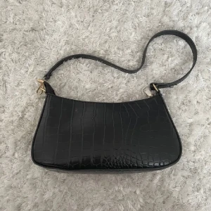 Svart axelväska i krokodilmönster - Snygg svart axelväska med krokodilmönster och guldfärgade detaljer. Perfekt för att ge en elegant touch till din outfit. Väskan har en justerbar axelrem och stängs med dragkedja.