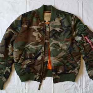 Camouflage bomberjacka - Säljer en cool grön camouflage bomberjacka från Alpha Industries. Jackan är mycket gott skick och har en klassisk bomberstil med dragkedja och ribbade muddar. Insidan är i en kontrasterande orange färg. Perfekt för en avslappnad stil. Jackan har använts av endast en person i ett fåtal gånger och kommer från ett djur- och rökfritt hem.