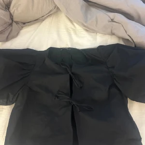 Svart blus med puffärmar och knytband - Elegant svart blus med puffiga kortärmar och knytband framtill. Perfekt för en stilren look.