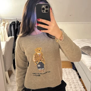 Beige stickad tröja från Ralph Lauren - Säljer en beige stickad tröja från Ralph Lauren med en ikonisk nallebrodyr på framsidan. Tröjan har långa ärmar och ribbade muddar vid ärmslut och nederkant. Perfekt för en avslappnad och stilren look.