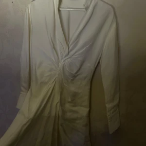 Vit omlottklänning från Sandro - Elegant vit omlottklänning med lång ärm och v-ringning. Klänningen har en stilren design med drapering framtill som ger en smickrande passform. Perfekt till studenten och sitter jättefint på kroppen och framhäver midjan 