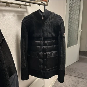 Svart stickad jacka från Moncler - Snygg svart stickad jacka från Moncler med quiltade detaljer och dragkedja framtill. Jackan har en hög krage och långa ärmar, perfekt för kyligare dagar. Den kombinerar stil och funktion med sina praktiska fickor.
