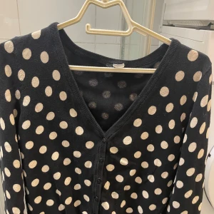 Svart kofta med prickigt mönster - Säljer en svart kofta med vita prickar. Koftan har en v-ringad design och knäppning framtill. Perfekt för att lägga till lite mönster i din garderob!