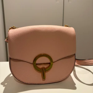 Rosa axelväska från Sandro med gulddetaljer - Rosa väska från Sandro med guldspänne framtill. Väskan har en justerbar axelrem och en kedja i guld. Insidan är klädd i en mjuk rosa mocka. Perfekt för att ge en touch av stil till din outfit. Väskan är endast använd ett fåtal gånger.