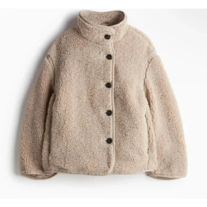 Beige teddyjacka med knappar - Mysig beige teddyjacka med hög krage och knäppning framtill. Perfekt för kyliga dagar med sin fluffiga och varma design. Jackan har en avslappnad passform och är ett stilrent tillskott till garderoben. Jackan är slutsåld på nätet och den är i bra skick