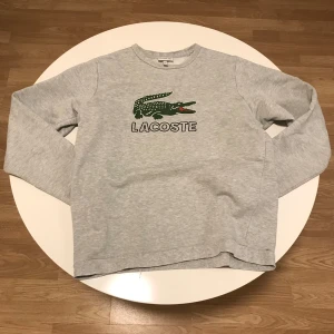 Grå tröja från Lacoste - Säljer en stilren grå tröja från Lacoste med det ikoniska krokodilmärket på bröstet. Tröjan har långa ärmar och en rund halsringning, perfekt för en avslappnad look. Ett litet hål nere vid midje vecket