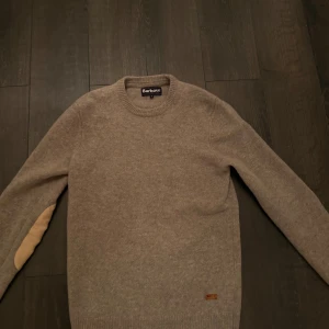 Beige tröja från Barbour - Säljer en stilren beige tröja från Barbour med rund hals och ribbade muddar. Tröjan har snygga armbågslappar i en ljusare nyans som ger en klassisk touch. Passar S