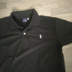 Svart pikétröja från Polo ralph lauren - Säljer en klassisk svart pikétröja från Polo ralph lauren i 100% bomull. Tröjan har en broderad logga på bröstet och knappar vid kragen. Perfekt för en stilren look.