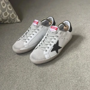 Vita sneakers med svart stjärna - Snygga vita sneakers med en svart stjärna på sidan och rosa detaljer på plösen. Skorna har snörning och en klassisk design som passar perfekt till vardags.