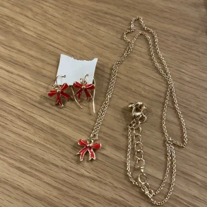 Guldhalsband och örhängen med röda rosetter - Charmigt set med ett guldhalsband och matchande örhängen, båda prydda med röda rosetter. Oanvända!