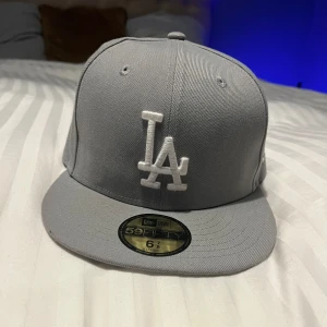 Grå keps från New Era - Snygg grå keps från New Era med broderad LA-logga framtill. Kepsen har en klassisk design med platt skärm och är perfekt för en casual look.