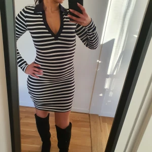 Klänning - Snygg randig klänning i svart och vit med en djup v-ringning och krage. Klänningen har en tight passform och långa ärmar, perfekt för en stilren look. Stl.xs