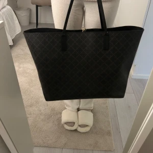 Svart shoppingväska från By Malene Birger - Snygg och rymlig svart shoppingväska från By Malene Birger med ett diskret rutigt mönster. Väskan har en praktisk innerficka med dragkedja för säker förvaring av småsaker. Perfekt för att bära med sig allt man behöver under dagen.