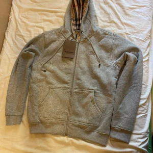 Grå hoodie från Burberry - Säljer en stilren grå hoodie från Burberry med klassiskt rutigt mönster i huvan. Tröjan har dragkedja framtill och praktiska fickor. Perfekt för en avslappnad och trendig look.