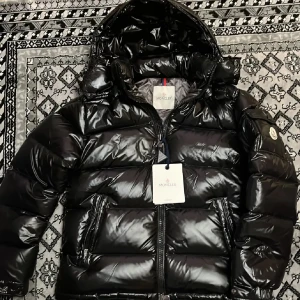 Svart pufferjacka från Moncler  - Snygg svart pufferjacka från Moncler med glansig finish och huva. Jackan har dragkedja framtill och två fickor med dragkedjor. Perfekt för kyliga dagar. Helt ny