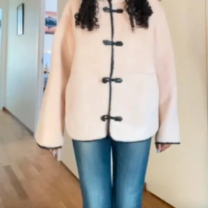 Beige teddyjacka med svarta detaljer - Säljer en mysig beige teddyjacka med svarta knappar och kantdetaljer. Jackan har en hög krage och långa ärmar, perfekt för kyligare dagar. Den har en loose passform som ger en avslappnad stil.