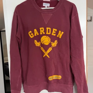 Vinröd tröja från GANT - Säljer en vinröd tröja från GANT med trycket 'GARDEN' i gult på framsidan. Tröjan har långa ärmar och en rund halsringning. Perfekt för en avslappnad stil.